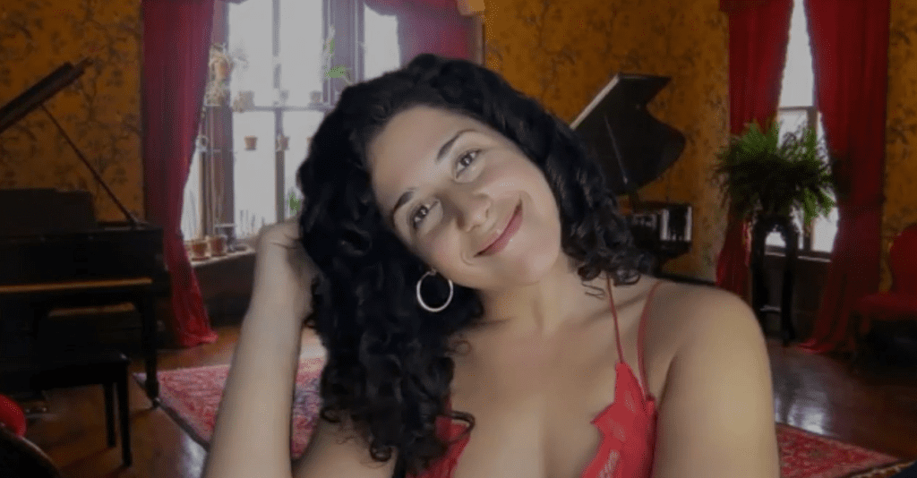A woman with curly hair smiles while tilting her head to the side in a warm and elegantly decorated room. She is wearing a red spaghetti-strap top and hoop earrings. She's role-playing in a murder mystery team building event.