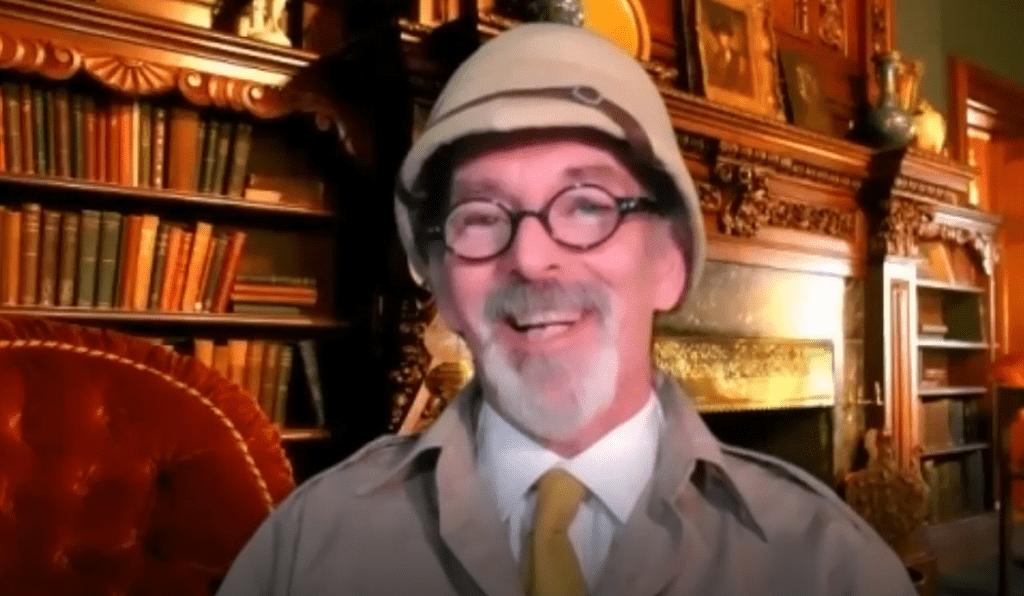 A man with glasses and a safari-style pith helmet smiles warmly in front of a cozy, vintage room filled with bookshelves. He is dressed in character, wearing a light-colored jacket and a yellow tie. The man is in character, part of a role-playing murder mystery team building event.
