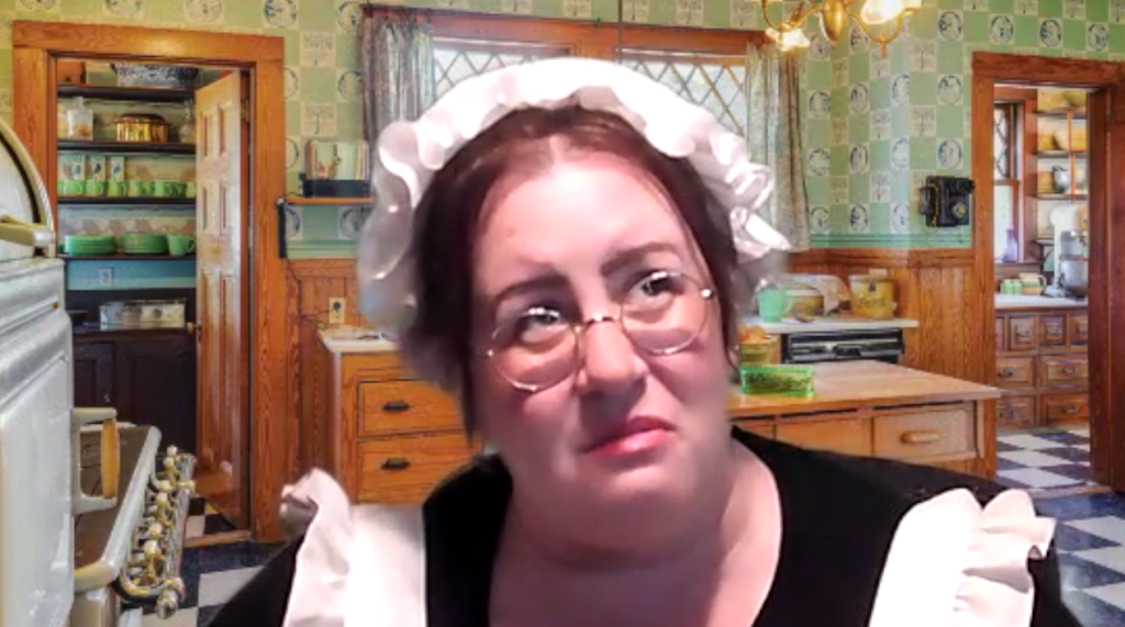 A woman dressed as a maid, wearing a white bonnet and glasses, is in an old-fashioned kitchen. She looks pensive or concerned as she gazes off to the side. The scene is part of a role-playing murder mystery.