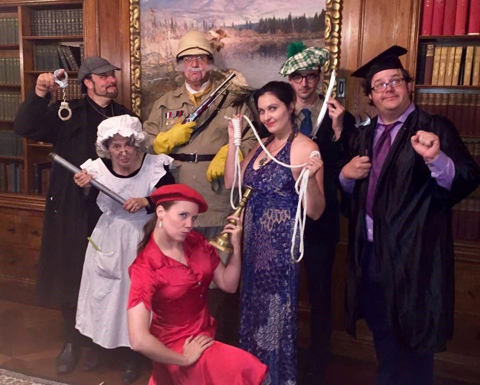 A group of seven participants pose in costume for a themed Murder Mystery Dinner team building event. Each person is dressed as a unique character, holding props that represent classic murder mystery items, such as a candlestick, rope, and revolver.