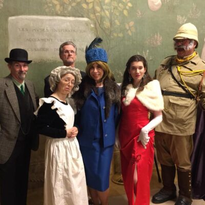 A group of actors dressed in period costumes for a Murder Mystery Dinner event. The characters include a man in a detective's coat and hat, a maid in a traditional white apron, a woman in a blue dress and fur hat, a glamorous woman in a red gown and fur shawl, and an explorer wearing a safari outfit and yellow gloves.