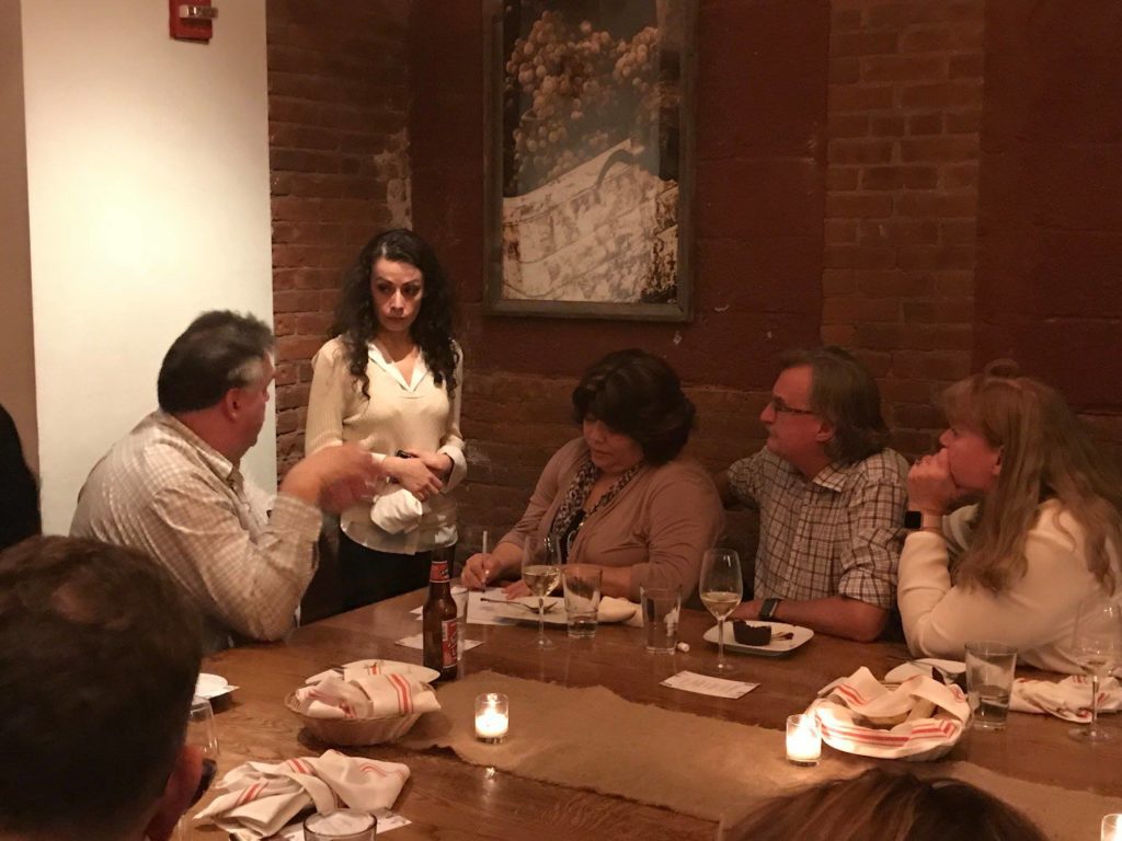 A group of four people sitting around a dimly lit dinner table engaged in conversation, with a female actor standing and looking at one of the seated individuals. The setting suggests an intimate dining experience, part of a Murder Mystery Dinner event, as the group interacts with the actor in the unfolding mystery.