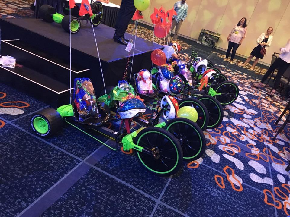 A row of assembled green Green Machine bikes is displayed on the floor, each adorned with colorful decorations, helmets, and balloons as part of a team building event.