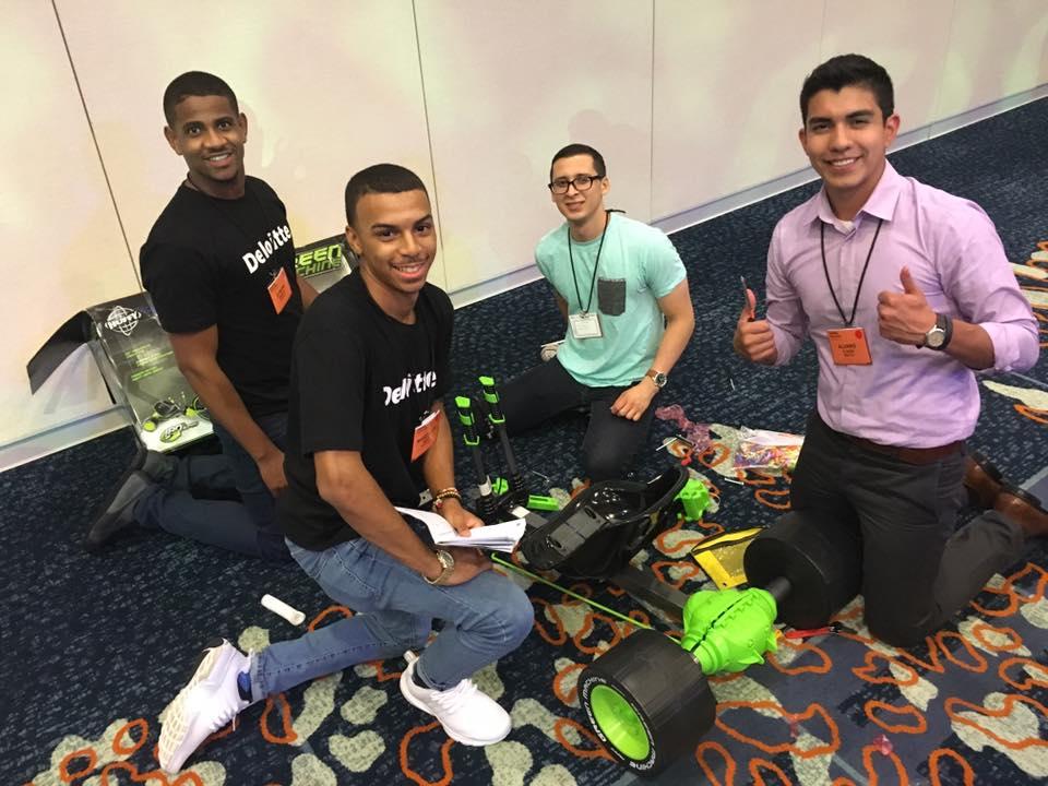 Four individuals kneeling on the floor assembling a Green Machine bike during a team building event. One person gives a thumbs-up while others smile, holding tools and instructions, as they work together on the project.
