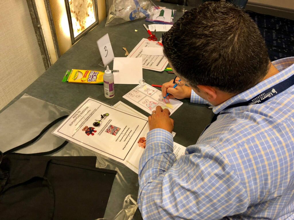 A man seated at a table is focused on drawing and coloring in a comic-style booklet. Various sheets of paper with superhero-themed drawings and instructions are spread out in front of him, along with a pack of colored markers and a bottle of glue. The scene captures creativity and concentration, as he works on a charitable team building activity.