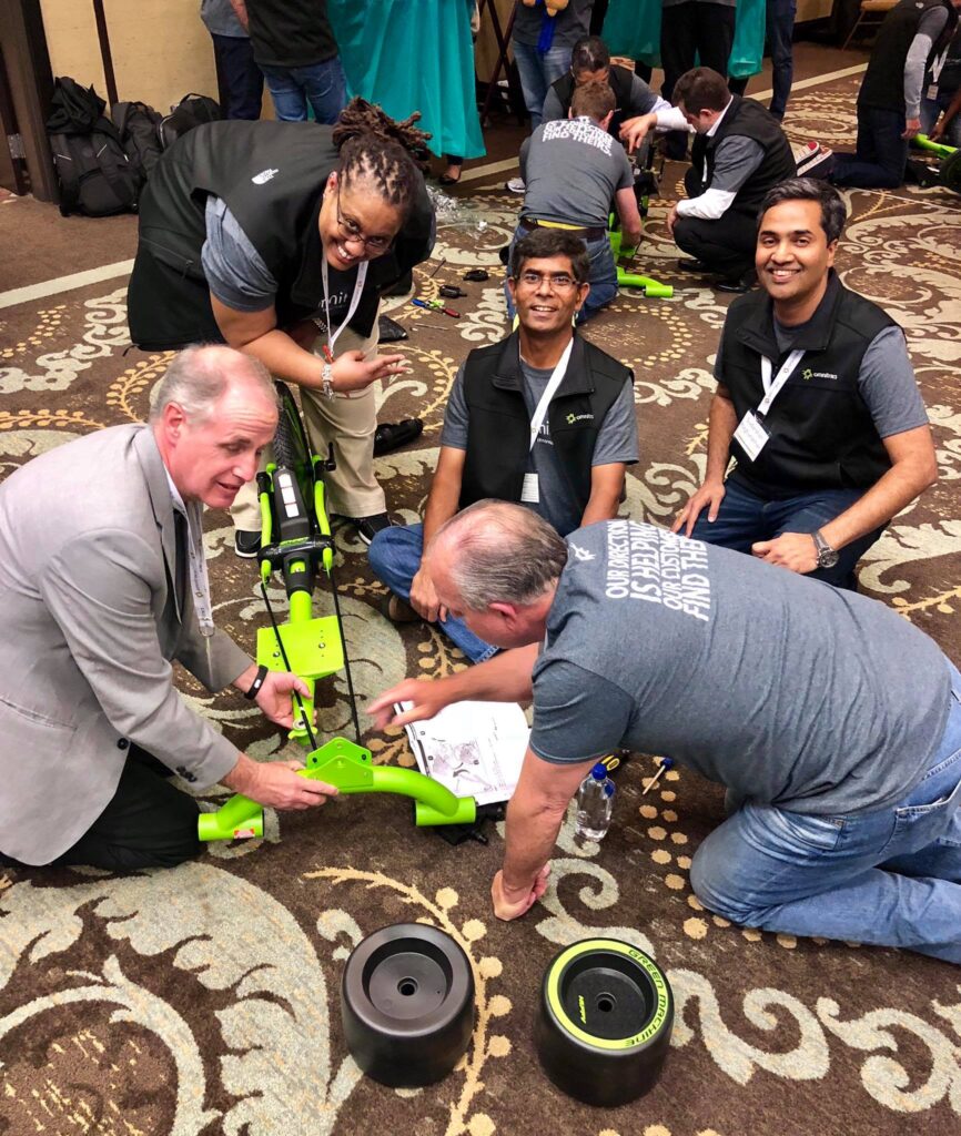A group of five people, some seated and others kneeling, work together to assemble a bright green bike. Two large wheels are placed on the floor next to the bike frame. The team appears to be engaged and smiling, collaborating on the project in a room filled with other teams working on similar bikes as part of a charitable team building activity.