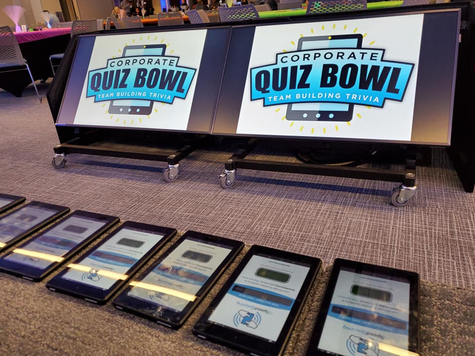 Corporate Quiz Bowl setup featuring large screens displaying the logo and a row of tablets ready for team participation in this engaging team building trivia event.