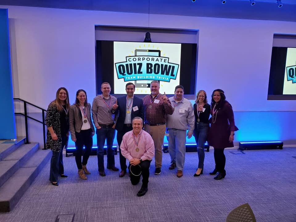 Smiling participants pose in front of a Corporate Quiz Bowl screen after completing a lively team building trivia competition.