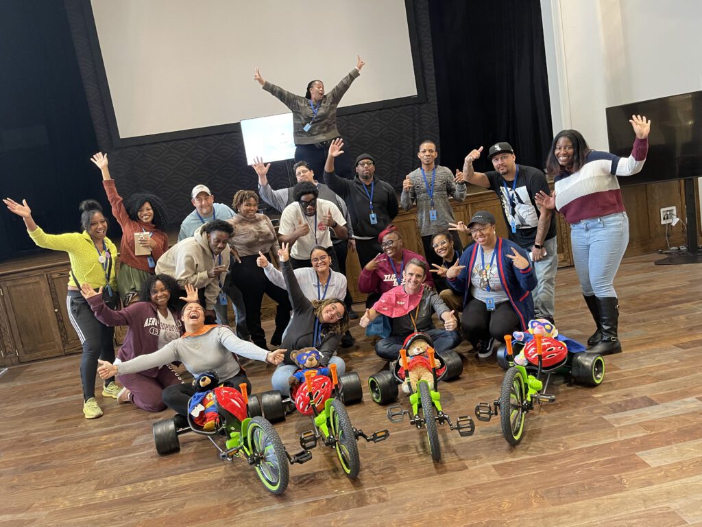 A group of people posing energetically with their arms raised, standing behind assembled Green Machine bikes. They are celebrating the accomplishment of a successful charitable team building event.