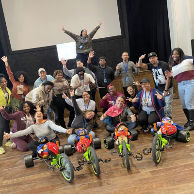 A group of people posing energetically with their arms raised, standing behind assembled Green Machine bikes. They are celebrating the accomplishment of a successful charitable team building event.