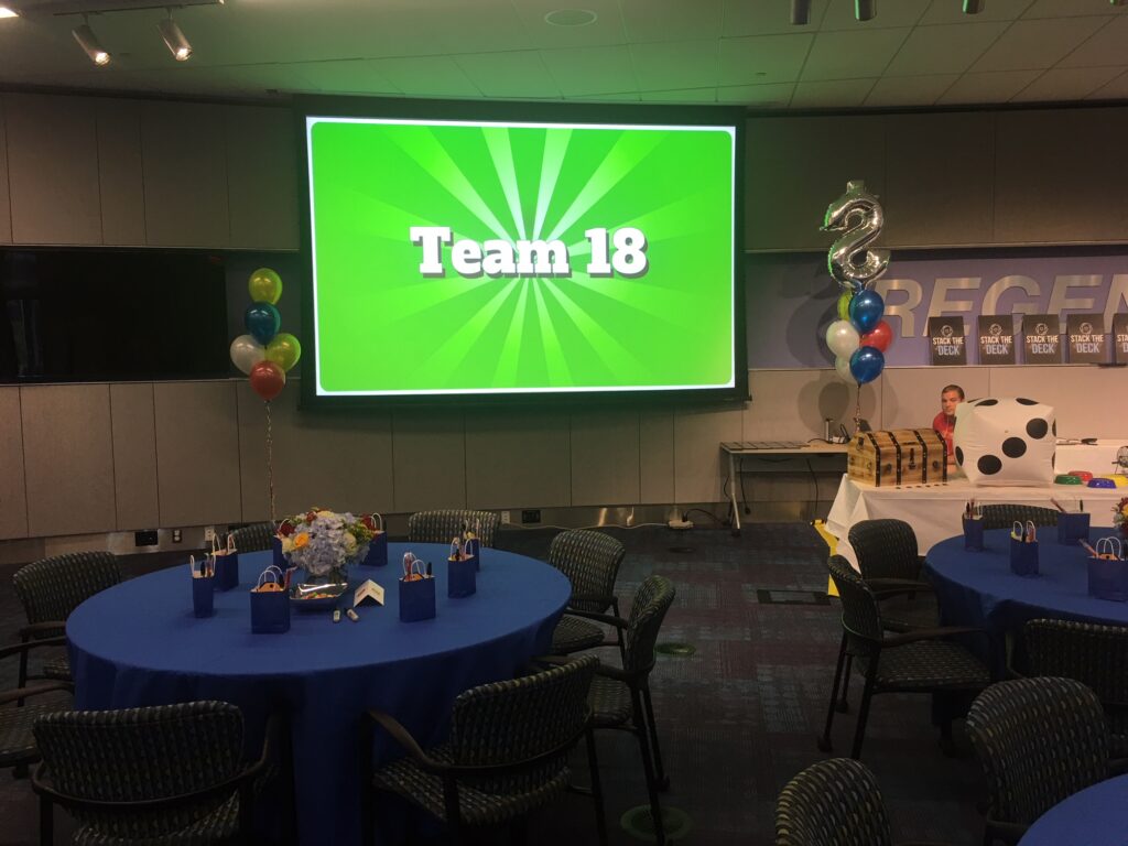 Corporate Quiz Bowl event with a large screen displaying 'Team 18', decorated tables with blue tablecloths, gift bags, balloons, and a playful game atmosphere.