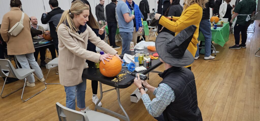 This image showcases a lively team building activity where participants are creating pumpkin racers. One person is working on carving or decorating a pumpkin, while another, wearing a festive witch's hat, appears to be using a power tool to attach components, possibly wheels, to the pumpkin. The setting has a group of people engaged in similar activities in the background, making it a fun and collaborative event, as part of a Halloween-themed competition.
