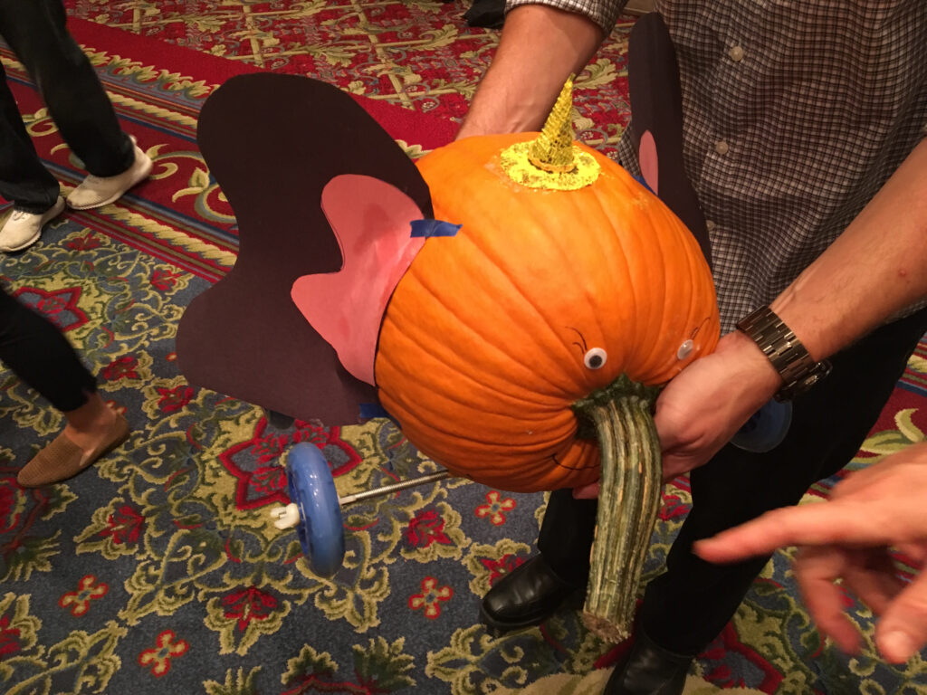 This image features a whimsical pumpkin racer designed to resemble an elephant. The pumpkin is decorated with large, pink and black ears attached to the sides and has a small horn or crown on top, adding a fun detail. The pumpkin’s stem is positioned to mimic an elephant's trunk, and googly eyes have been added for an expressive touch. The racer is equipped with wheels, ready for the competition. The colorful, patterned carpet background adds to the playful atmosphere of this creative team building event.