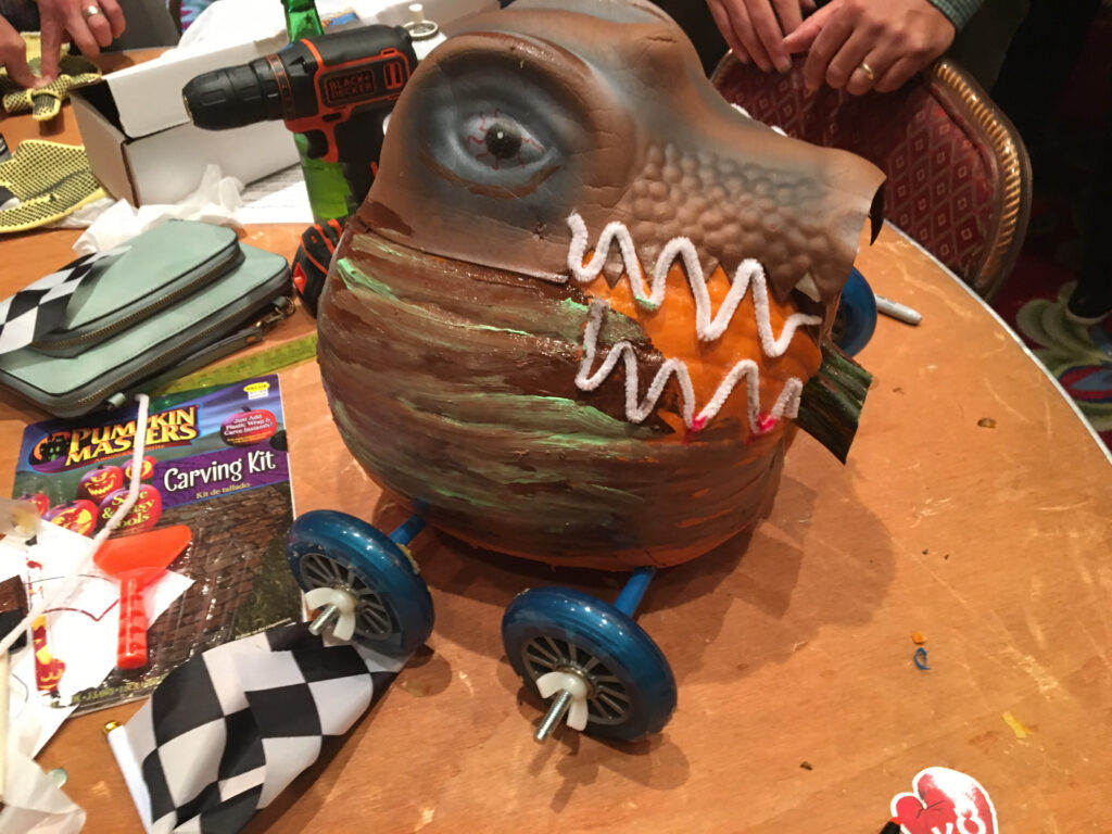 This image showcases a creatively designed pumpkin racer. The pumpkin has been intricately transformed to resemble a fierce creature, possibly inspired by a dinosaur or dragon, with painted scales and a set of sharp teeth. The design is very detailed, with wheels attached to the bottom for racing. A power drill and a pumpkin carving kit are visible in the background, suggesting that the team has been hard at work on this impressive creation.