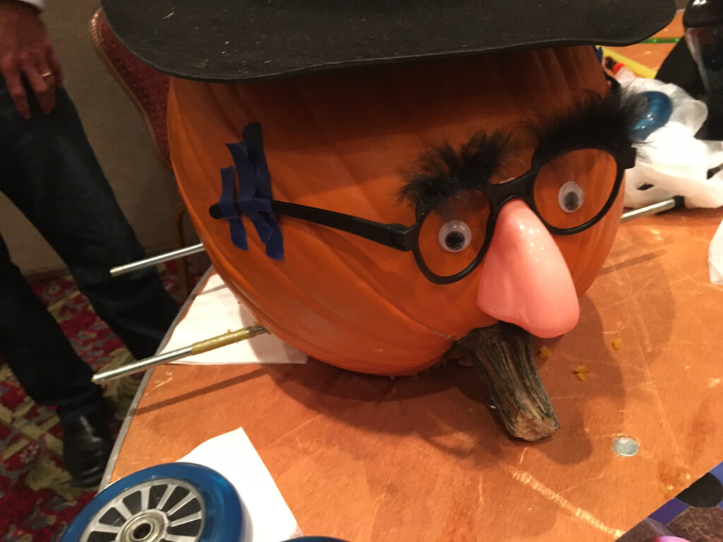 This image shows a creatively decorated pumpkin racer featuring a humorous face. The pumpkin has been outfitted with a comical disguise: a fake nose, glasses, and bushy eyebrows, giving it a playful personality. Axles for the wheels are visible, suggesting that this pumpkin is ready to be part of the pumpkin race. The setup adds a whimsical element to the team building activity, blending creativity with competition.