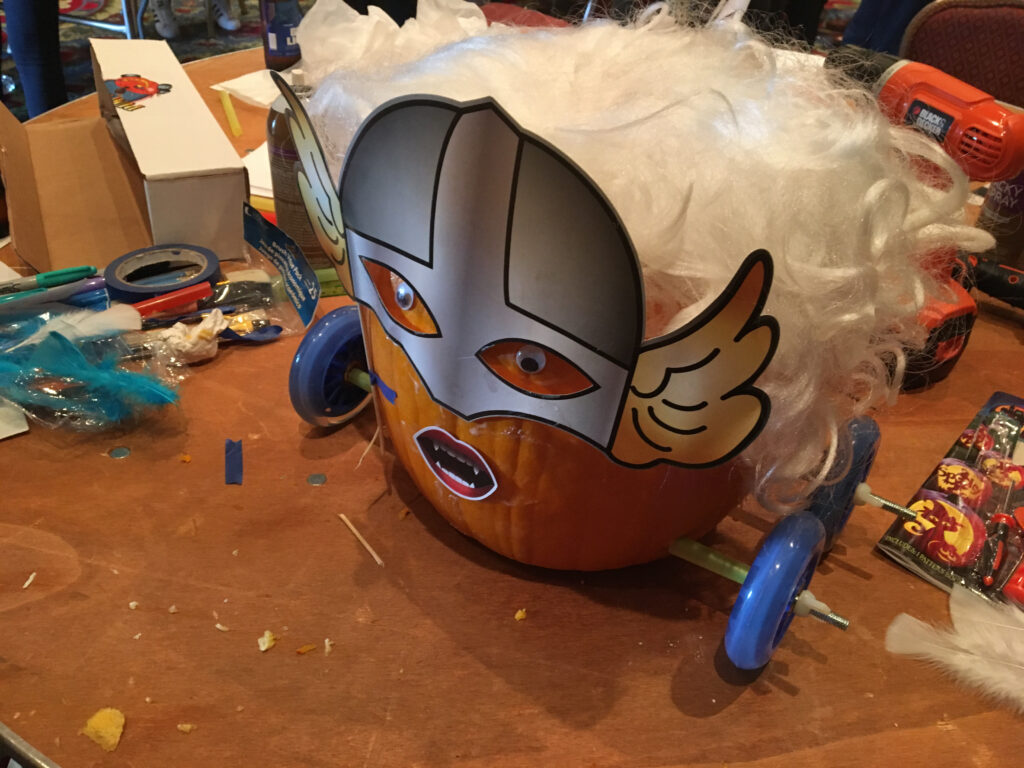This image showcases a creatively designed pumpkin racer. The pumpkin has been transformed with a Viking-style mask, featuring wings on the side and dramatic facial expressions. The pumpkin is adorned with a fluffy white wig, adding a playful element to the design. The racer is fitted with blue wheels, and the tools and materials scattered around the workbench suggest that the team is in the midst of finalizing their creation. This fun and imaginative design emphasizes the lighthearted competition of the pumpkin race team building event.