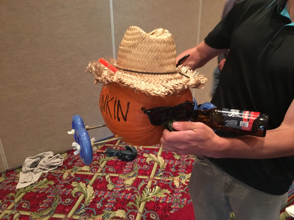 This image shows a creatively designed pumpkin racer, styled with personality. The pumpkin is adorned with a straw hat and sunglasses, giving it a cool, laid-back look. One of the wheels appears to be part of a toy or plastic assembly, attached to make the pumpkin mobile. The pumpkin also seems to be holding a bottle, adding to the humorous, festive vibe. It’s a fun and engaging way to bring creativity and teamwork to a pumpkin racing team building event.