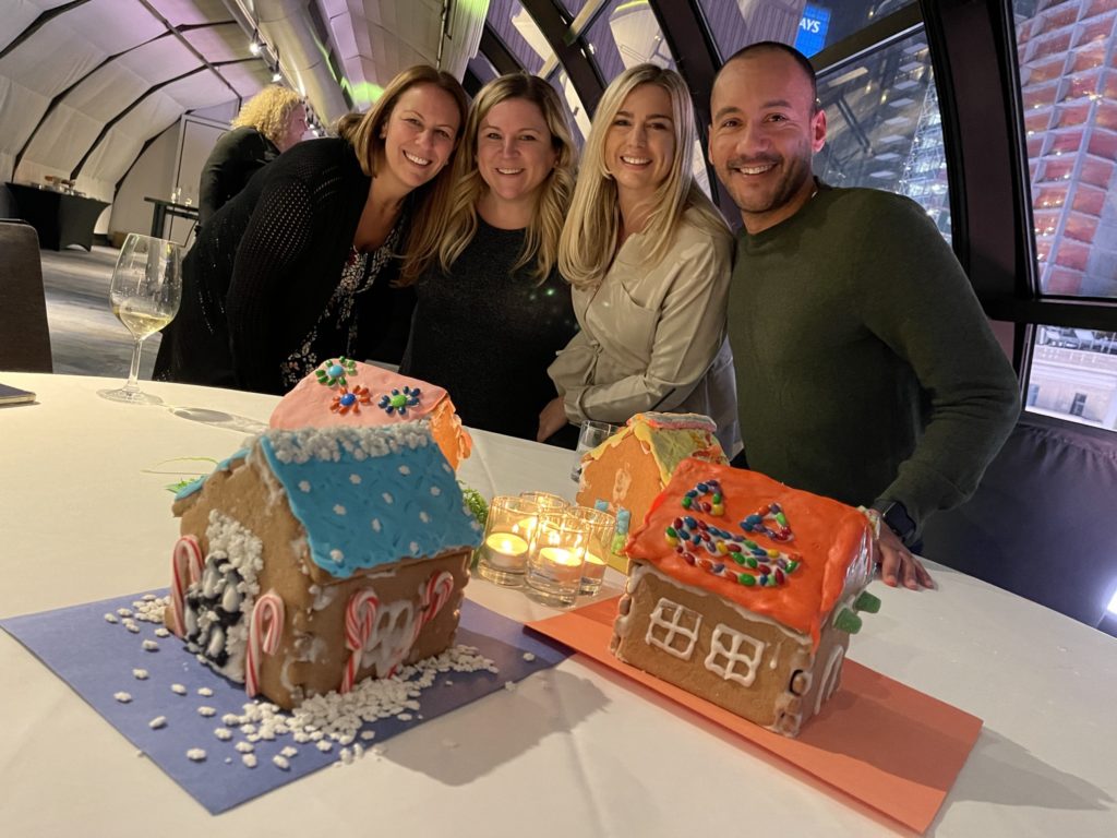 A group of four participants smiling and standing behind a table displaying three colorfully decorated gingerbread houses during a gingerbread house team building competition. The table is decorated with small lit candles, adding a cozy touch to the festive atmosphere.