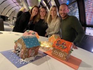 A group of four participants smiling and standing behind a table displaying three colorfully decorated gingerbread houses during a gingerbread house team building competition. The table is decorated with small lit candles, adding a cozy touch to the festive atmosphere.