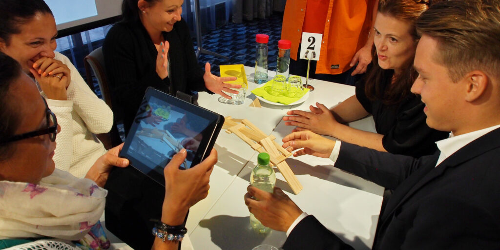 A team gathers around an iPad while engaging in a hands-on challenge, working together to complete a task in the Quickfire team building event.