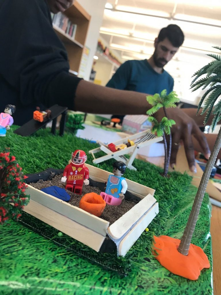 A close-up of a miniature playground scene at a City Build team building event, featuring toy figurines, a sandbox, palm trees, and vibrant greenery, with participants working in the background.