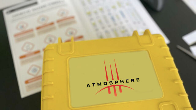 Close-up of a yellow case labeled 'Atmosphere' used in a team building activity, with instruction sheets visible in the background.