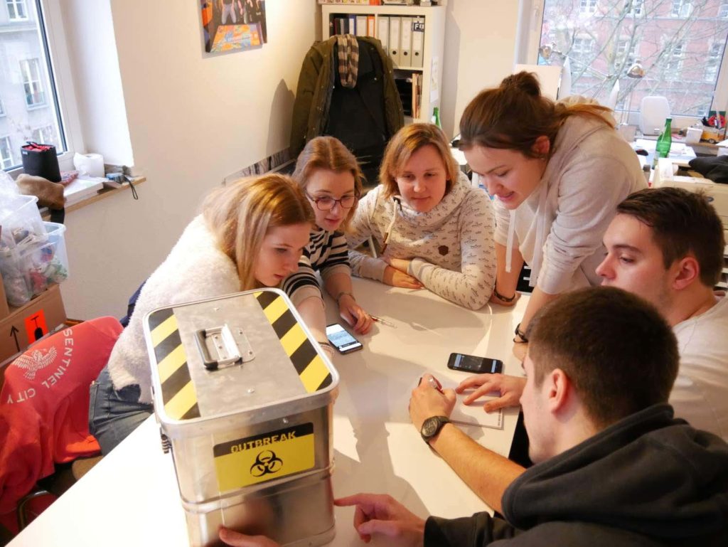 Team collaborating to solve a puzzle around a biohazard-themed box during the 'Atmosphere' team building challenge.