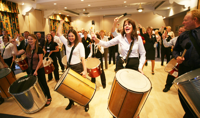 A group of energetic participants playing large percussion drums during a lively indoor BeatsWork team building session. They are smiling and engaged, enjoying the rhythm and teamwork experience.