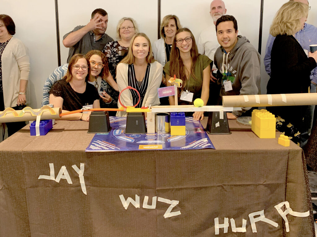Team members gather proudly around their completed chain reaction machine at a team building event, featuring creative structures and labeled with playful text on the table.