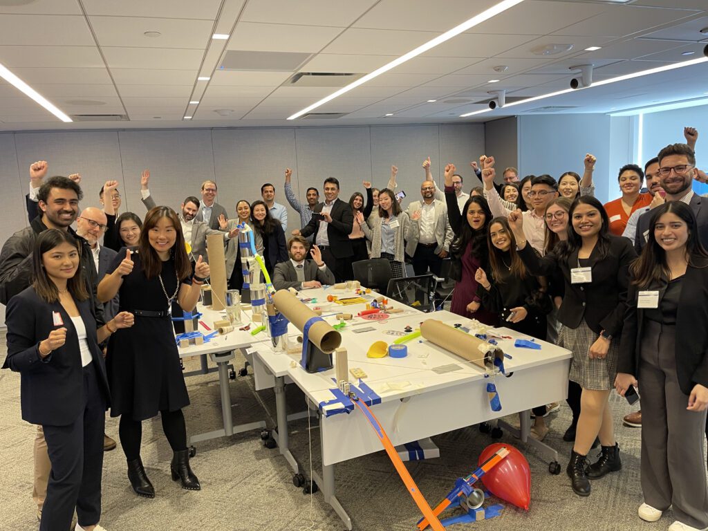 Group of participants celebrate the completion of their collaborative Chain Reaction machine during a team building event, gathered around a complex setup of interconnected parts on a table.