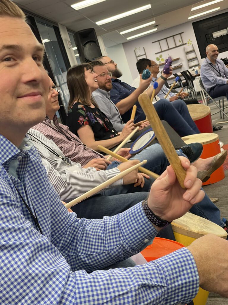 Participants seated in a circle, practicing rhythm on percussion instruments during a BeatsWork team building activity.