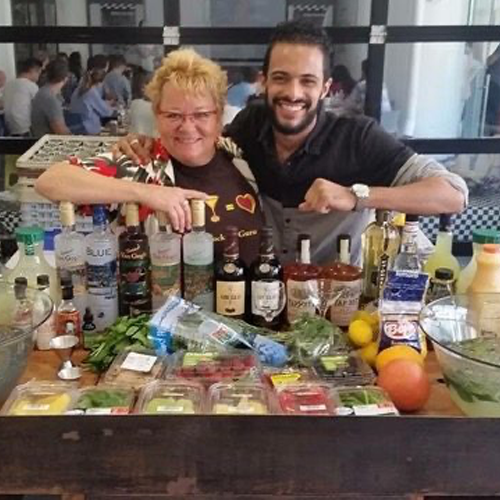 Two participants posing happily behind a bar setup filled with various bottles, mixers, and fresh ingredients during the Team Mixology team building event, excited to learn cocktail-making skills.