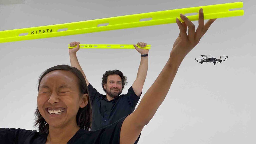 Participants lift obstacle bars with excitement as a drone flies by during the Game of Drones team building challenge, emphasizing fun and teamwork.