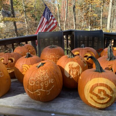 A collection of intricately carved pumpkins displayed outdoors on a wooden table during a team building pumpkin carving event. The pumpkins feature various logos, designs, and creative carvings, with an American flag in the background, set against a scenic autumn forest backdrop.