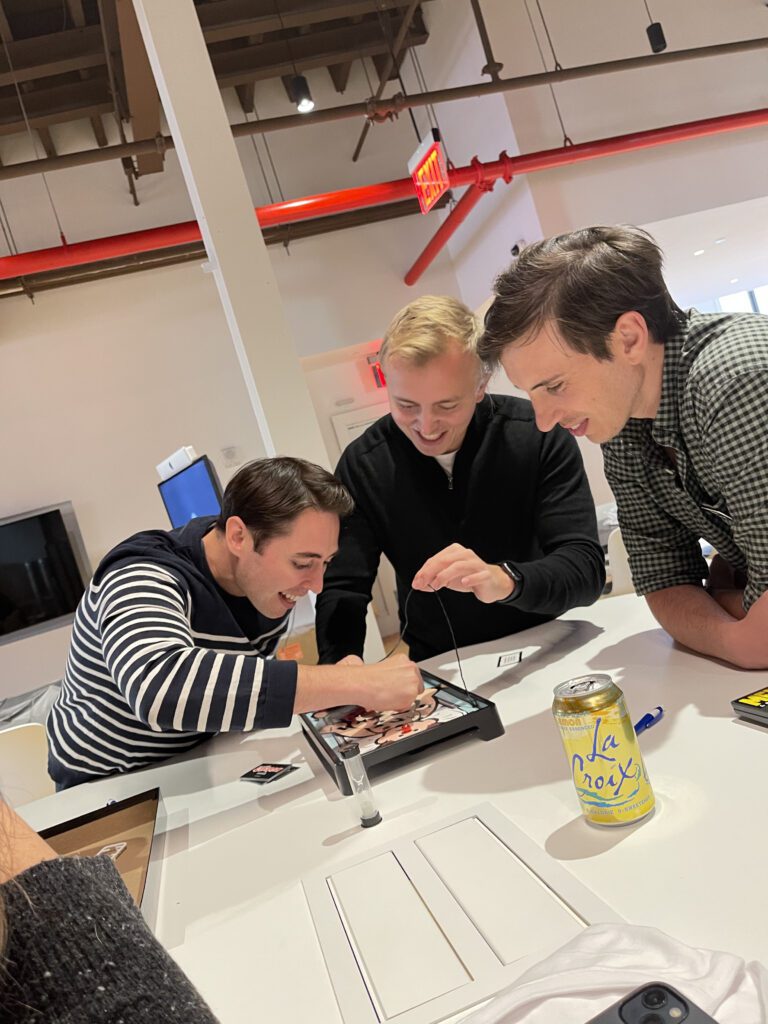 Three participants enthusiastically play the game 'Operation' at a table during the Helping Hands team building event. They are laughing and focused on completing the challenge to earn supplies for care kits.