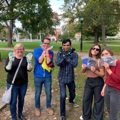 A group of five participants is standing outside in a park, each holding up socks and care items collected for a Healthcare Heroes scavenger hunt team building challenge.