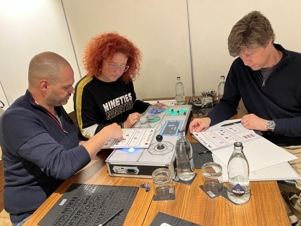 Three team members focus on the Team Synergy retro gaming console, analyzing instructions and working together to solve mini-game challenges during a team building event.