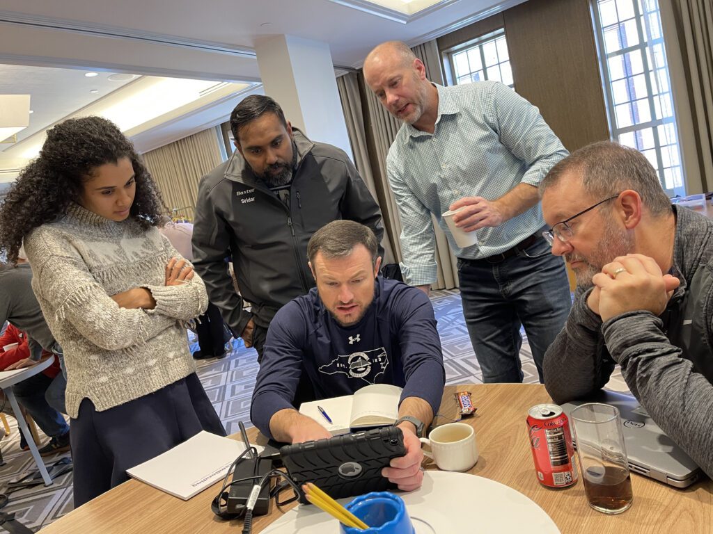 A group of five team members gathered around a tablet, focusing on completing a task during the Wheels Around the World team building event.