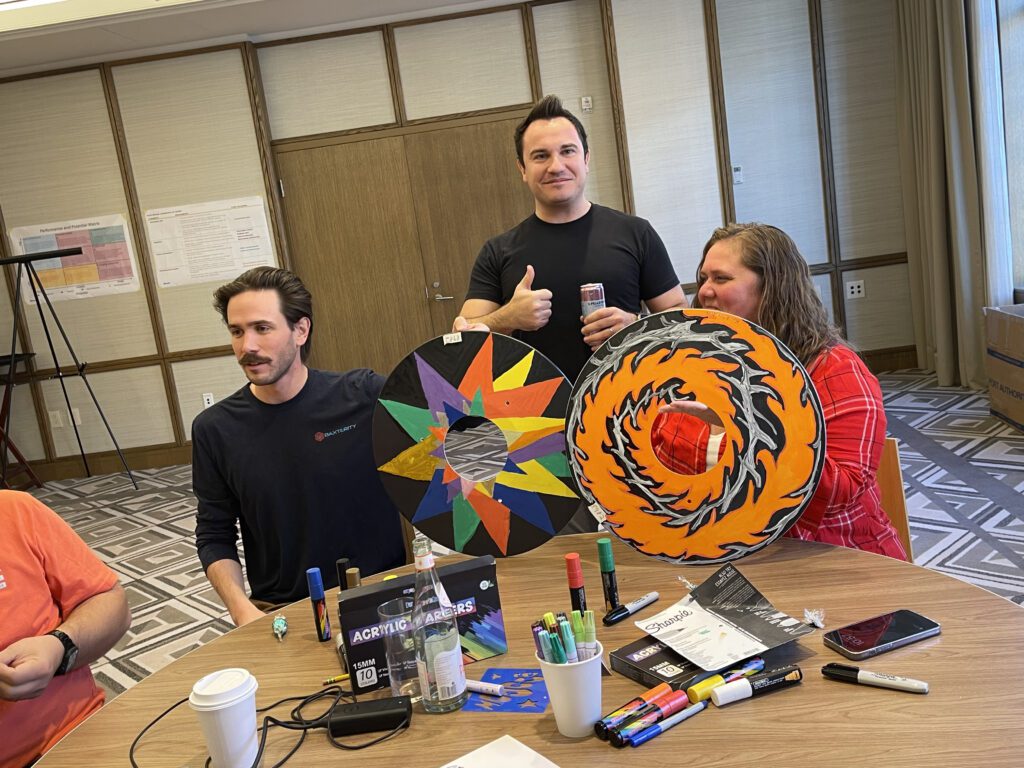 Three participants in the Wheels Around the World team building event display their creatively decorated wheelchair safety covers. The covers feature vibrant designs, showcasing the team's artistic contribution to the charitable project.
