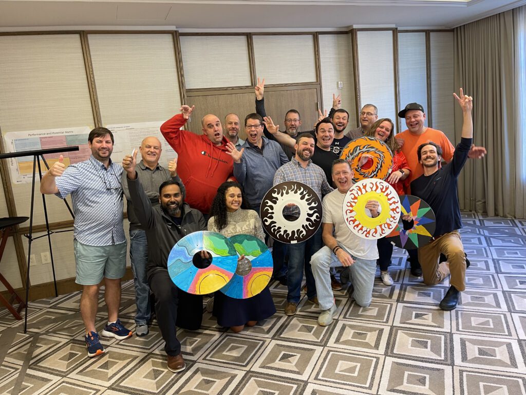 A group of enthusiastic participants proudly displaying personalized wheelchair safety covers they decorated during the Wheels Around the World team building event. The decorated wheels feature colorful designs, and the group celebrates their contribution to making a positive impact.