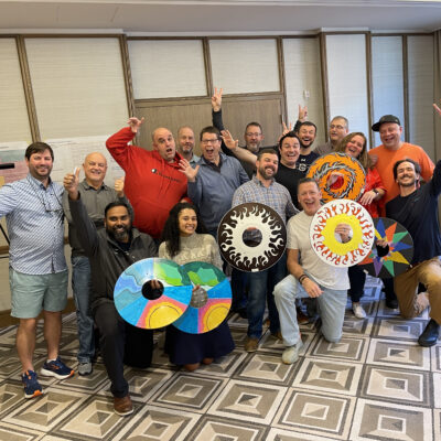 A group of enthusiastic participants proudly displaying personalized wheelchair safety covers they decorated during the Wheels Around the World team building event. The decorated wheels feature colorful designs, and the group celebrates their contribution to making a positive impact.