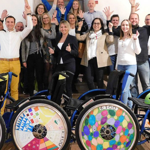 Team members stand behind two completed and decorated wheelchairs, smiling and raising their hands, celebrating their efforts in the Wheels Around the World team building event.