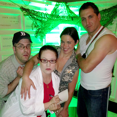 Four individuals dressed in costume pose for a group photo in front of a green-lit backdrop. The person on the far left is wearing a baseball cap and glasses, with a playful, surprised expression. Next to them, a person in a white lab coat with glasses holds a notepad, looking serious. The third person, wearing a patterned top, smiles confidently while resting an arm on the shoulder of the person on the far right, who is wearing a white tank top and strikes a tough pose with arms crossed.