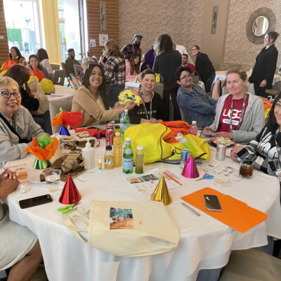 A group of people seated around a table, smiling and interacting, with colorful party hats, birthday-themed decorations, and supplies spread out, engaging in a team building activity for a charitable cause.