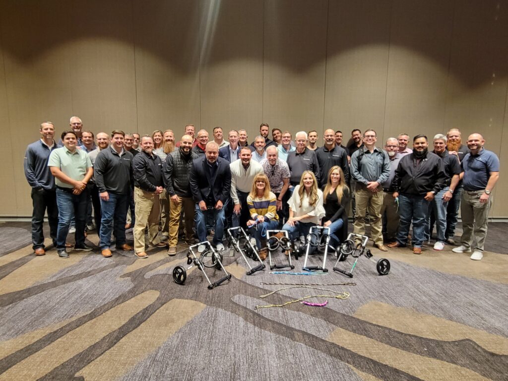 A large group of participants proudly standing together after successfully assembling pet wheelchairs during a team building event, showcasing their completed projects in front of them. The focus is on teamwork and making a meaningful impact on animal welfare.
