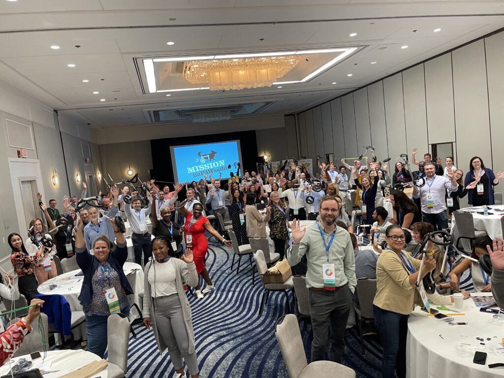 A large group of participants enthusiastically wave during a Mission ImPAWssible team building event, with tables set up for assembling pet wheelchairs in the background.