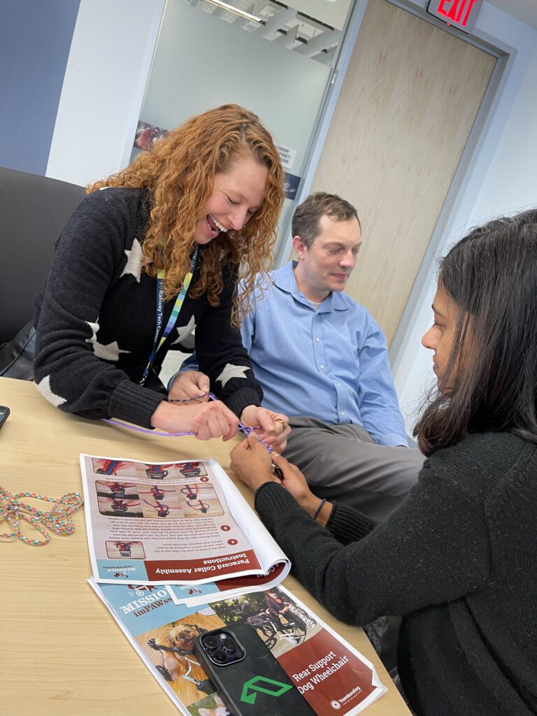 Three team members working together, smiling as they assemble components for a pet wheelchair. They focus on tying ropes while a Mission ImPAWssible brochure is on the table. This scene highlights teamwork and collaboration in creating life-changing mobility aids for pets in need.