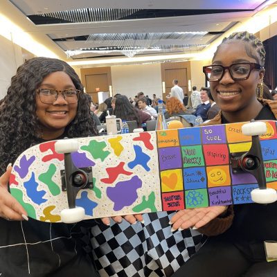 Two participants proudly display their colorful and creatively decorated skateboard, made during the Just Roll With It charitable team building event.