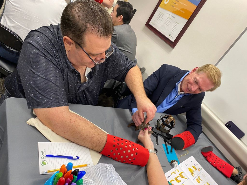 Two participants are actively assembling a prosthetic hand during The Prosthetic Hand Project. They are wearing red prosthetic simulation sleeves, adding a level of challenge and empathy to the task. One person is holding and connecting the components of the mechanical hand while the other assists. A variety of tools and assembly instructions are visible on the table, and a notebook and colorful pens are seen nearby, indicating careful planning and attention to detail. This image highlights the teamwork, focus, and effort involved in assembling prosthetic hands for donation to those in need.