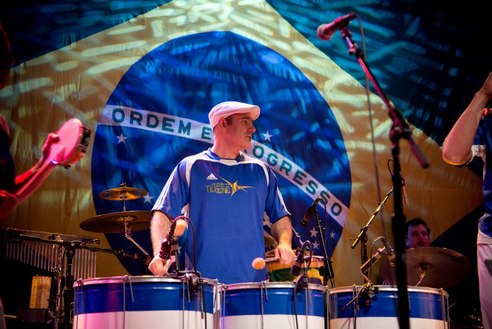 Keynote speaker Josh Robinson performs energetically on Brazilian drums in front of a large Brazilian flag backdrop. Wearing a blue jersey and white cap, Josh leads an engaging musical performance.